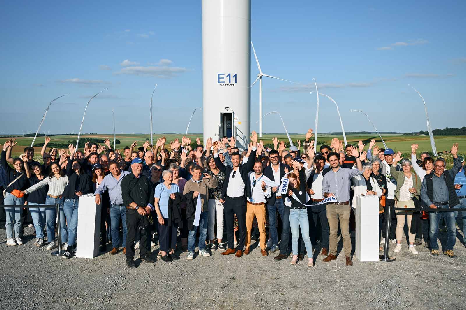 Un grand groupe de personnes se tenant devant une éolienne, souriant et agitant la main avec un ciel clair en arrière-plan.