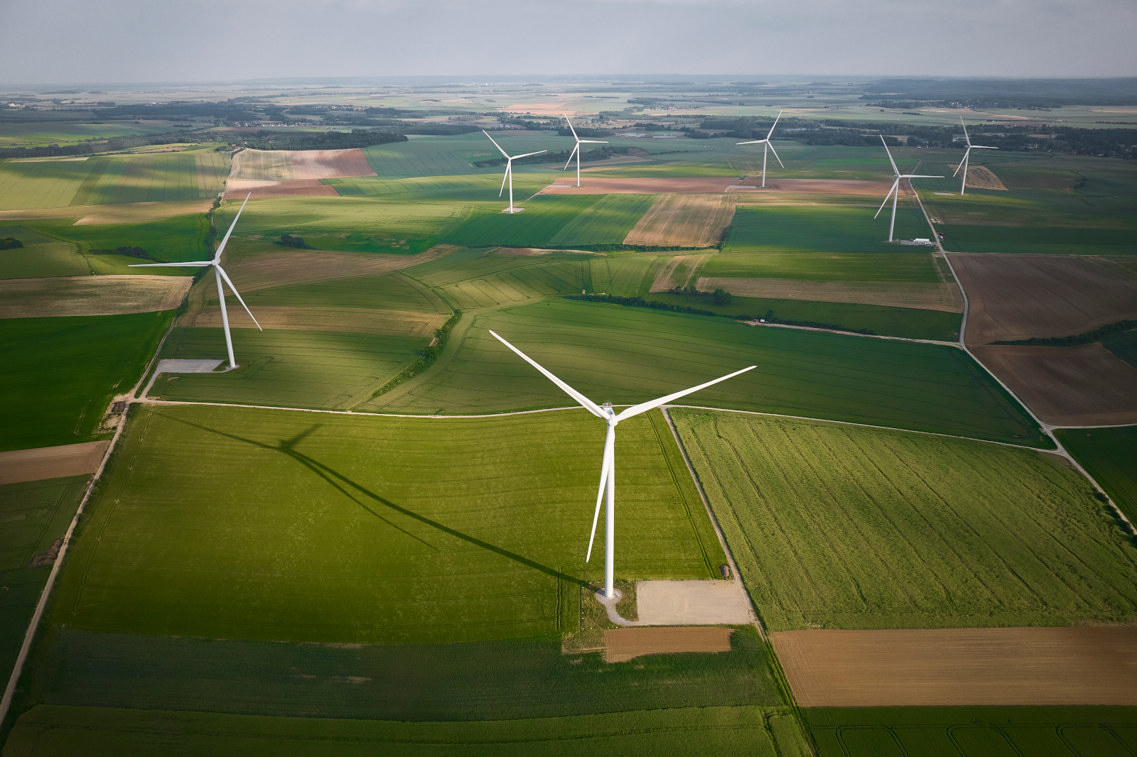 Aerial view of green fields with multiple wind turbines casting long shadows across the landscape under a cloudy sky.