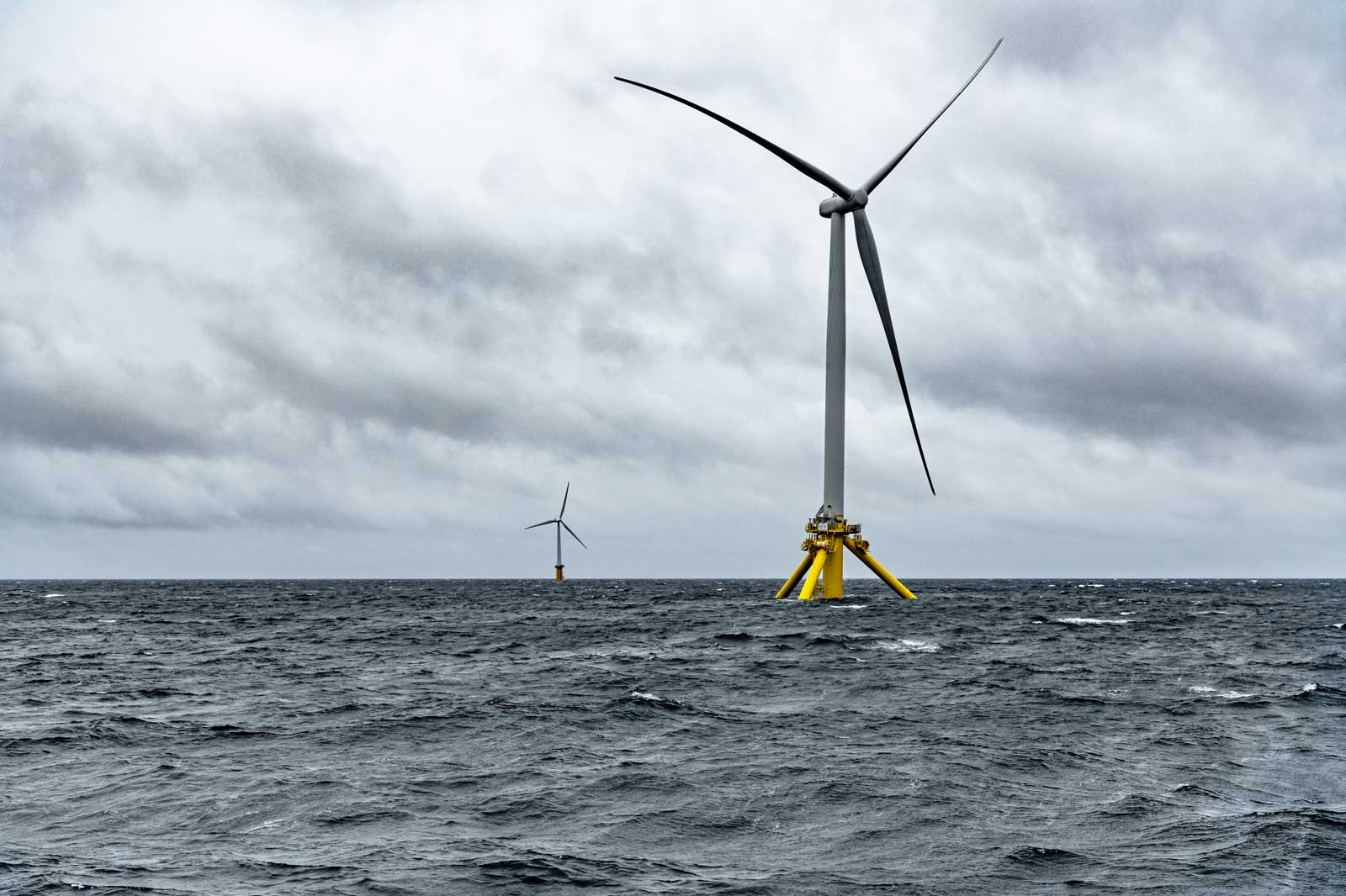 Two offshore wind turbines stand in turbulent sea beneath a cloudy sky.