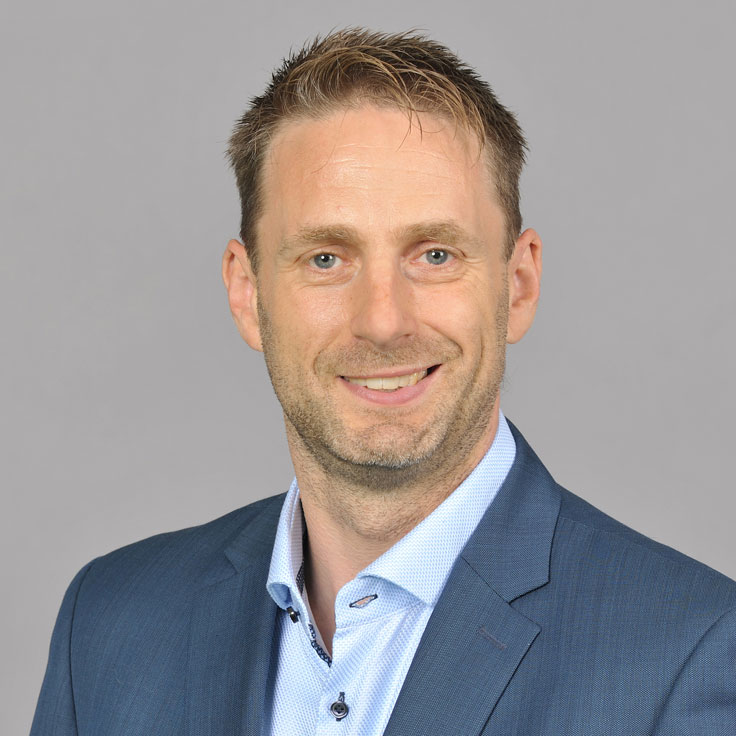 A man is wearing a blue suit and a light blue shirt against a grey background. Portrait of Matthias Beigel.