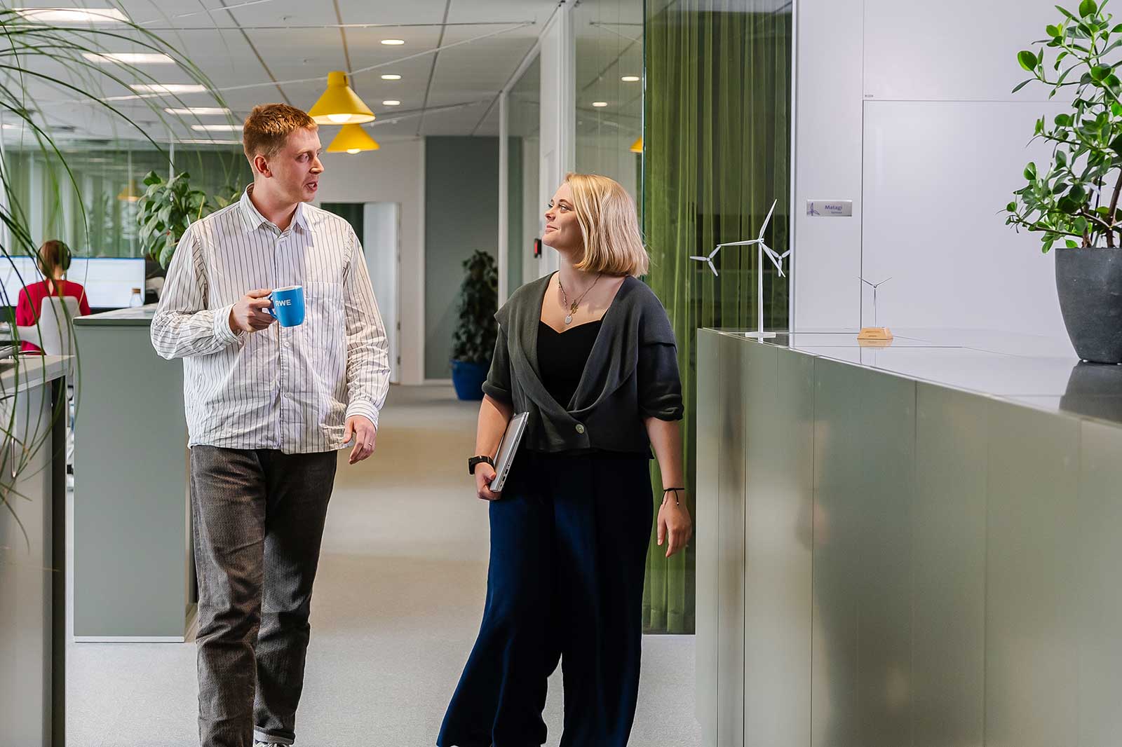 A man and a woman walk in a modern office, surrounded by plants and a wind turbine model.