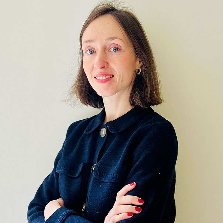 Une personne avec les cheveux bruns courts portant une veste noire à col, les bras croisés, sur un fond uni.