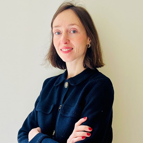 A person with short brown hair wearing a black collared jacket, arms crossed, against a plain background.