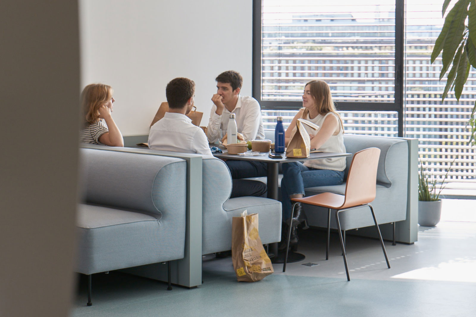 A group of four people sit at a round table in a bright, modern space, enjoying food and drinks together.