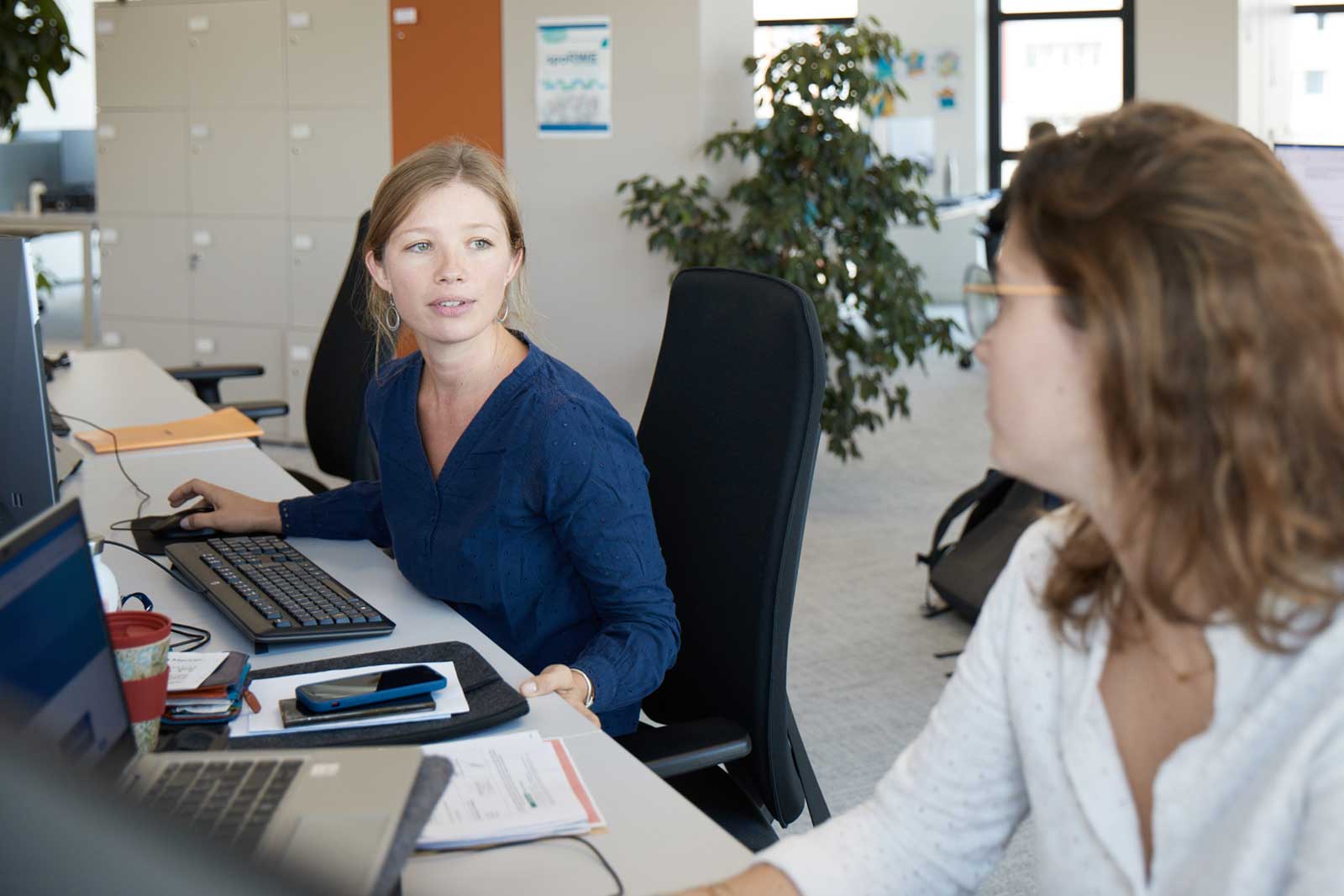 Une femme en blouse bleue est assise à un bureau avec des ordinateurs, des papiers et une tasse à proximité, dans un environnement de bureau lumineux.