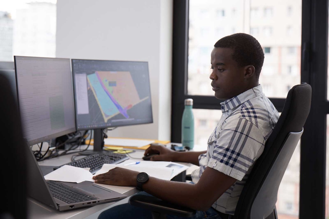 A man seated at a desk with multiple monitors, using a laptop and surrounded by documents.