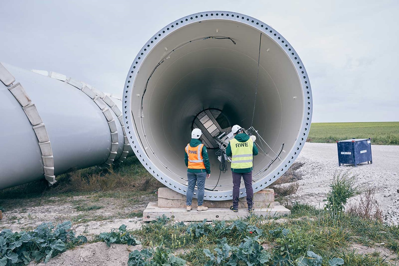 Two workers inspect a large wind turbine section, wearing safety gear, with green fields in the background.