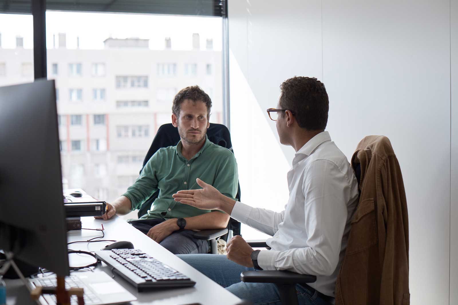 Two men discuss at an office desk, with monitors and a window providing a city view in the background.