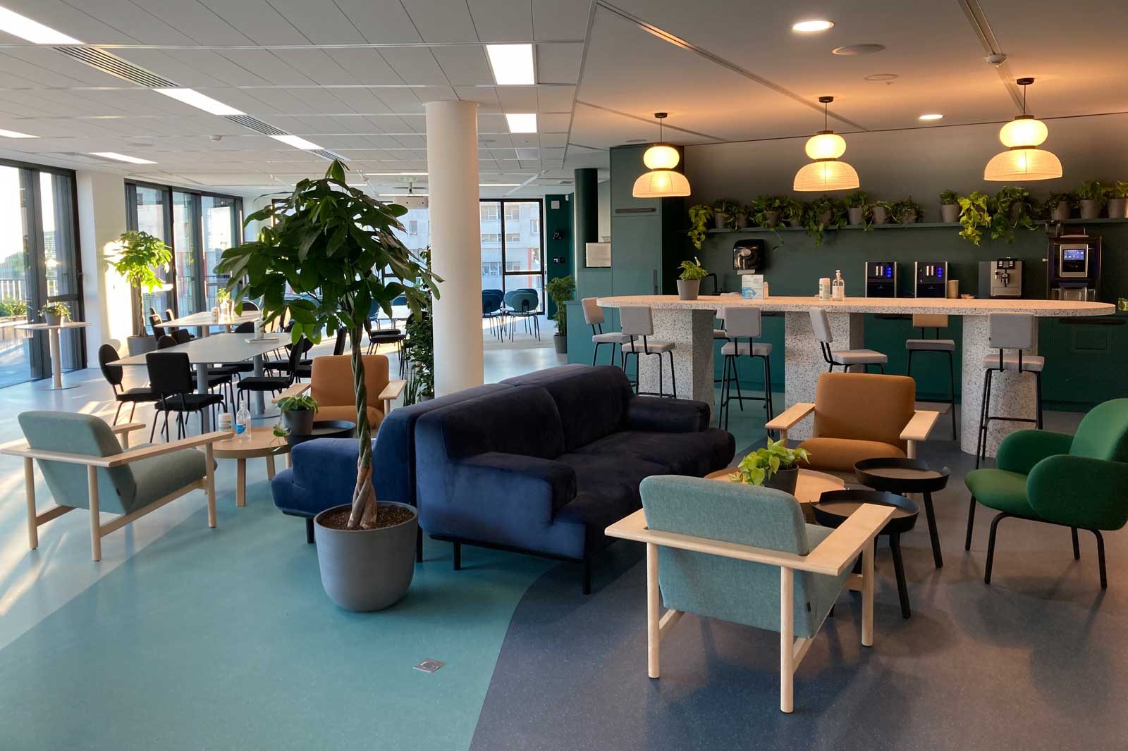 A modern lounge featuring plants, seating areas, and a bar. Bright colours and ample daylight create an inviting atmosphere.